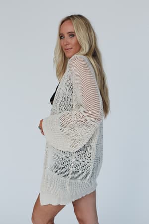 A woman with long blonde hair models the Brenden Knit Netted Cardigan in Ivory over a black outfit, standing against a plain light background and glancing over her shoulder at the camera.