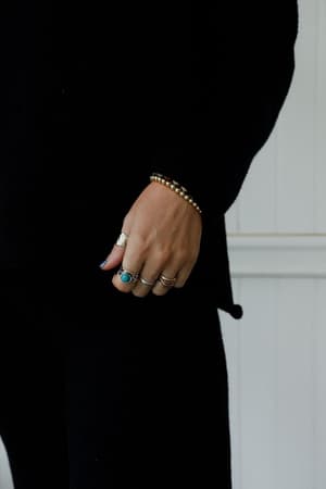 A person in a black long-sleeve outfit displays their left hand with several rings, including the Kapalua Bay Adjustable Turquoise Ring - Silver and boho jewelry, plus a gold beaded bracelet, against a white paneled background.