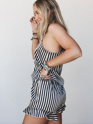 A smiling model shot in profile wearing a black and white striped satin short set with a ruffled hem, layered with stacked beaded bracelets and rings — playful and confident against a clean light gray backdrop.