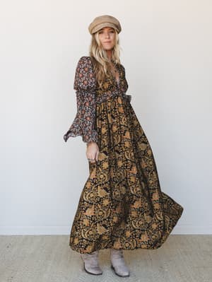 A flowy black maxi dress with golden floral and paisley print catches mid-movement, styled with a tan baker boy cap and grey ankle boots for an effortlessly boho, autumn-ready look.