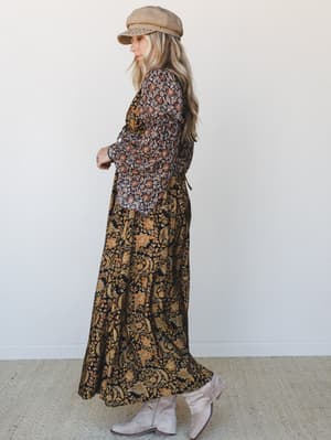 A flowy mixed-print maxi dress in deep black and golden florals, styled with suede ankle boots and a studded newsboy cap — the kind of outfit that looks like it was pulled together effortlessly but absolutely wasn't.