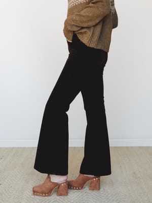 A side-profile shot of velvet flare trousers in a deep chocolate brown, styled with a chunky earth-tone sweater and studded suede platform clogs — the kind of outfit that makes a Tuesday feel intentional.