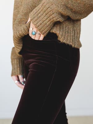A close-up detail shot of deep burgundy velvet trousers paired with an olive marl ribbed sweater, hands tucked into the pocket and stacked with turquoise and gold rings — earthy, collected, and quietly rich in texture.