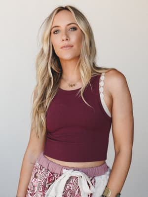 A relaxed, confident close-up of a model in a deep burgundy ribbed crop tank layered over a lace bralette, paired with a paisley-print skirt — the kind of effortless mix that looks thrown together in the best way.