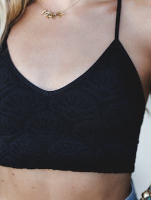 A close-up detail shot of a black lace-texture bralette with thin straps and a soft v-neckline, styled with a gold 'MAMA' letter necklace — relaxed, personal, and quietly expressive.