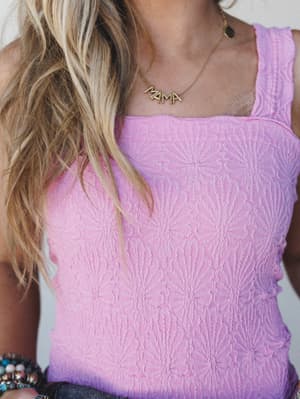 A close-up of a lavender-pink textured tank top with a tonal floral jacquard pattern, styled with denim and a gold 'MAMA' letter necklace — the kind of top that does the work for you.