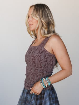 A side-profile shot of a model in a fitted mauve-brown textured tank with a tonal floral jacquard pattern, styled with a plaid skirt and a stack of turquoise and cream boho bracelets — the kind of outfit that looks like it came together effortlessly