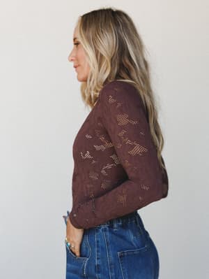 A side-profile shot of a long-sleeve lace bodysuit in deep burgundy, styled with high-waist denim and a turquoise ring — the kind of top that quietly does a lot.