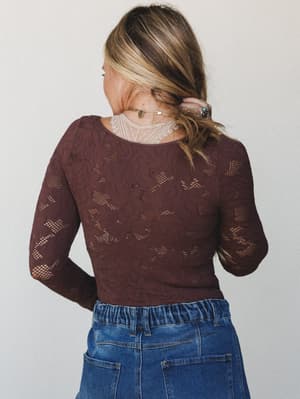 Shot from behind, a model in a deep brown lace bodysuit and medium-wash jeans tucks a loose wave behind her ear — the open lace pattern and gold layered necklaces doing all the quiet storytelling.