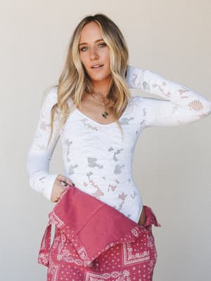 A long-sleeve white lace bodysuit with a floral cutout pattern is styled casually with a rose-pink bandana print skirt tied at the waist, giving the whole look that effortless, thrown-together-on-purpose energy.