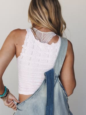 A back-view shot showing a soft icy pink lace-back cami layered under light wash denim overalls, styled with stacked boho bracelets — casual and a little dreamy.