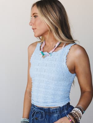 A sun-warmed model shot in profile wearing a textured periwinkle blue cropped cami layered with a colorful beaded ribbon necklace and stacked bracelets — easy, summery, and just the right amount of put-together.