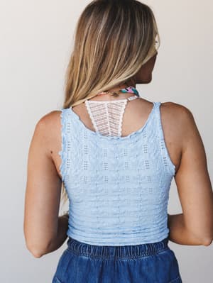 A back-view shot of a cropped knit cami in soft sky blue with a lace panel cutout at the upper back, paired with a denim skirt — casual and a little sweet, like something you'd grab last-minute and still look put-together.