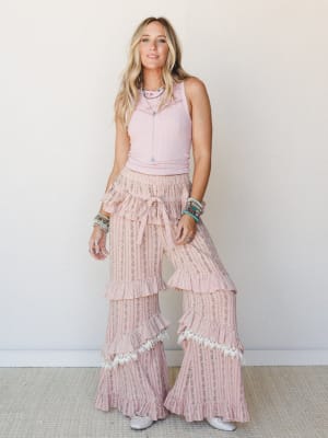 A sun-kissed blonde model in a blush ribbed crop tank and dreamy wide-leg ruffle pants with lace trim and a floral stripe print — the whole look reads like a country-festival love letter in pink.