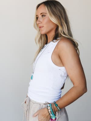 A side-profile shot of a model in a white ribbed tank with subtle lace detailing, styled with linen-look pants and layered boho jewelry — the kind of effortless warm-weather look that works as hard as you don't want to.