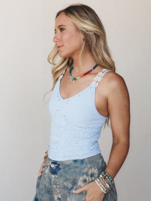 A sun-kissed model in profile wears a soft periwinkle blue ribbed cami with delicate eyelet strap detail, layered with a multicolor beaded necklace and stacked bracelets — the whole look has that easy, collected summer energy.