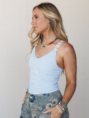 A sun-kissed model in profile wears a soft periwinkle blue ribbed cami with delicate eyelet strap detail, layered with a multicolor beaded necklace and stacked bracelets — the whole look has that easy, collected summer energy.