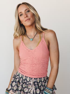 A relaxed close-up of a model in a textured pink cami with pointelle and cable-knit detailing, styled with a floral print skirt, stacked bracelets, and a turquoise butterfly necklace — the kind of outfit that looks effortless but clearly wasn't an ac
