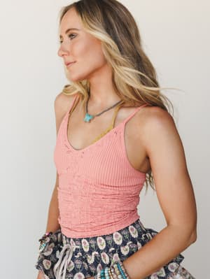 A sun-kissed model shot in profile wearing a coral-pink ribbed cami tucked into a navy floral skirt, styled with turquoise jewelry and stacked beaded bracelets — the kind of effortless summer look that feels like it came together naturally.