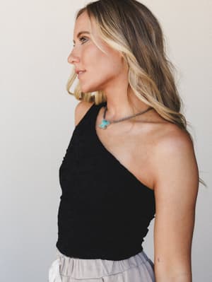 A profile shot of a model in a fitted black one-shoulder brami top with smocked texture, paired with a tiered light gray skirt and a turquoise butterfly pendant necklace — effortlessly cool with a hint of boho.