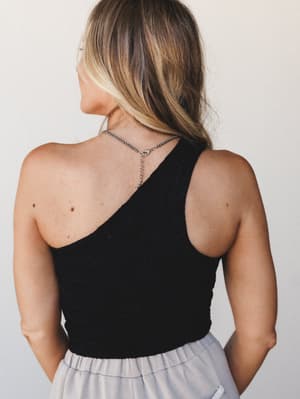 A back-facing shot of a textured black one-shoulder brami top paired with a soft lavender skirt, styled with a delicate silver bead necklace — effortless and a little unexpected.