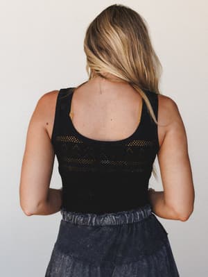 A back-facing shot of a model in a black open-knit tank bodysuit with a geometric pointelle pattern, paired with a charcoal acid-wash skirt — relaxed and a little editorial at once.