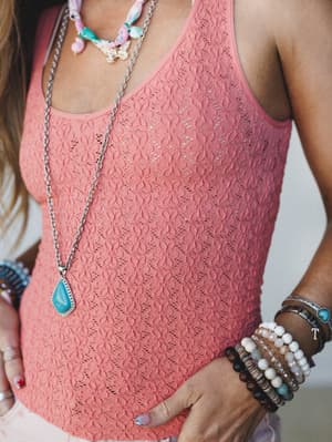 A close-up torso shot of a coral pointelle tank styled with layered boho jewelry — turquoise stone pendant, butterfly charm necklace, and a full stack of beaded bracelets that feel like they were collected one trip at a time.