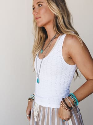 A sun-kissed model shot in profile wearing a white crochet-knit brami tank layered with stacked turquoise jewelry and beaded bracelets, paired with a striped skirt — the whole look reads like a slow Saturday somewhere warm.