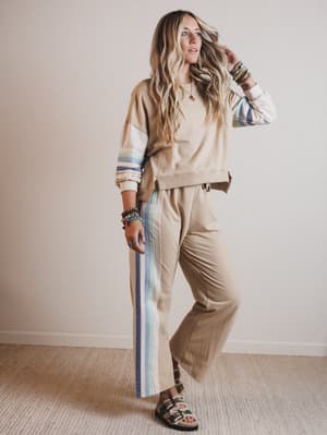 Woman wearing a beige sweater with colorful sleeves and striped pants against a plain wall.