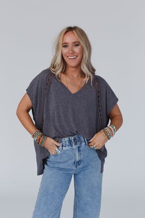 A woman with long blonde hair smiles at the camera, wearing The Nest Camden Lace V Neck Top in Charcoal, high-waisted blue jeans, layered necklaces, and multiple colorful beaded bracelets on both wrists.