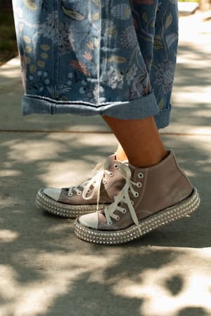Rocking the Chantel Studded High Top Sneaker in Mocha, this person completes their playful boho look with rhinestone-studded sneakers, wide white laces, and blue floral jeans with rolled-up cuffs on a sunlit sidewalk.