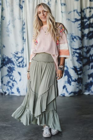 A woman with long blonde hair smiles by a blue and white curtain, wearing the Washed Ashore Ruffled Maxi Skirt in olive, a light pink embroidered top, stacked bracelets, necklaces, and white sneakers for a festival chic look.