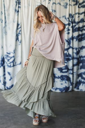 A woman stands indoors wearing a loose lavender top, platform sandals, and the Washed Ashore Ruffled Maxi Skirt in Olive. She looks down with her hand in her hair, set against a blue and white tie-dye curtain for a festival-chic vibe.