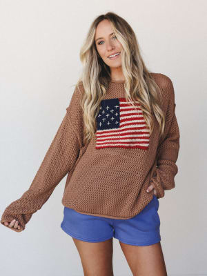 An oversized open-knit sweater in warm camel with a chunky American flag graphic on the chest, paired with periwinkle mini shorts — casual and a little nostalgic, like a summer cookout that turned into a bonfire.