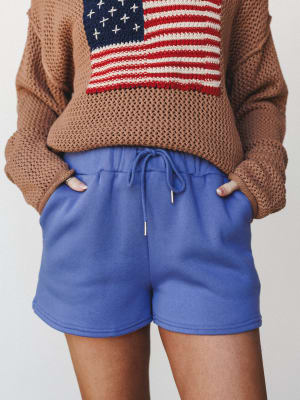 A cozy, Americana-leaning outfit pairing periwinkle blue sweat shorts with a camel open-knit sweater featuring an embroidered American flag graphic — relaxed, expressive, and effortlessly worn-in.