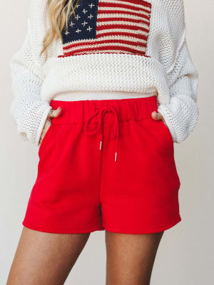 A mid-shot of red drawstring shorts paired with a cream chunky knit sweater featuring an American flag graphic — casual, playful, and ready for a backyard cookout or a lazy Fourth of July morning.