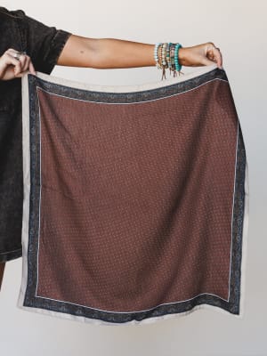 A model holds up a large square scarf displaying a rust brown ditsy floral print with a navy paisley border and cream edge — stacked turquoise and beaded bracelets adding that effortless boho layering moment.