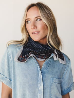 A close-up model shot of a woman in a light wash chambray button-down styled with a navy and rust bandana scarf tied loosely at the neck — easy, expressive, and effortlessly pulled-together.