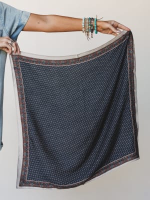 A model holds out a navy and cream square scarf with a floral border print, her stacked turquoise and beaded bracelets adding a laid-back boho finishing touch against a soft neutral studio backdrop.