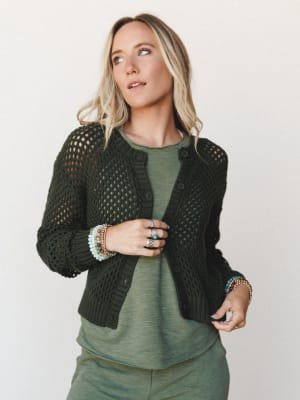 A model layers an open-knit dark olive cardigan over a sage green tee, stacked with beaded bracelets and silver rings — the kind of effortless tonal dressing that looks intentional without trying too hard.