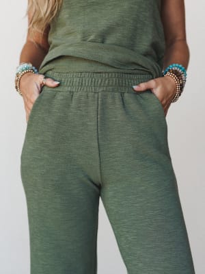 A close mid-shot of olive wide-leg pants with a smocked elastic waistband, styled as part of a matching set, with stacked beaded bracelets adding a relaxed boho touch.