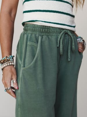 A close-up of wide-leg sweat pants in washed sage green styled with a cream and dark green striped textured top, accessorized with stacked beaded bracelets and silver rings — relaxed and put-together at the same time.
