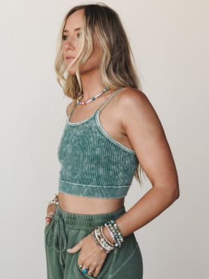 A side-profile shot of a model in a washed ribbed crop cami and drawstring jogger pants in matching sage-to-forest green, layered with colorful beaded jewelry that gives the whole look a relaxed, collected feel.