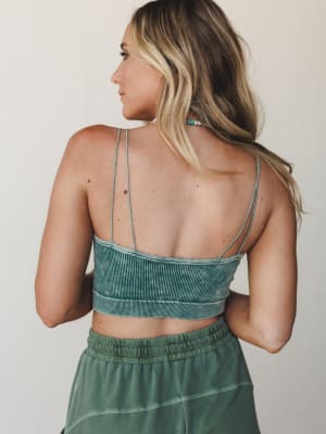 A back-view shot showing the strappy open-back detail of a ribbed crop bralette top in washed sage green, paired with a matching elastic-waist skirt — effortless and summery with a boho-casual ease.