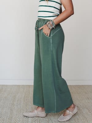 A side-profile shot of wide-leg fleece pants in washed dark green, styled with a striped cream-and-green crop top, woven flats, and a stack of beaded bracelets — relaxed and put-together at the same time.