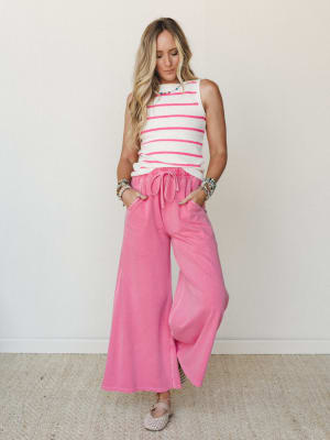 A bright, playful outfit pairing wide-leg pink drawstring pants with a cream and hot pink striped sleeveless sweater top, styled with stacked beaded bracelets and woven flats against a clean white studio wall.