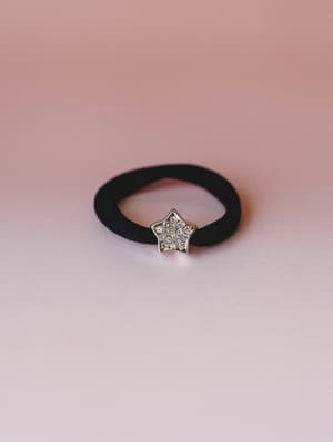 A black elastic hair tie centered with a rhinestone-studded gold star charm, shot against a soft blush gradient — small but the kind of detail that makes a ponytail feel intentional.