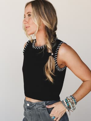 A side-profile shot of a model in a fitted black crop tank with white whipstitch trim at the neckline and shoulders, paired with grey washed denim and stacked boho bracelets — effortless and a little editorial.