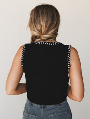 Shot from behind, a model in a black sleeveless cropped top with white whipstitch trim along the neckline and armholes pairs it with grey denim — simple, considered, and quietly cool.