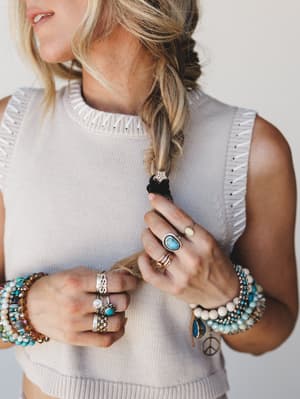 A close-up lifestyle shot focused on layered boho jewelry — stacked beaded bracelets, turquoise stone rings, silver bands, and a peace charm — styled against a cream sleeveless knit crop top with whipstitch trim detail.